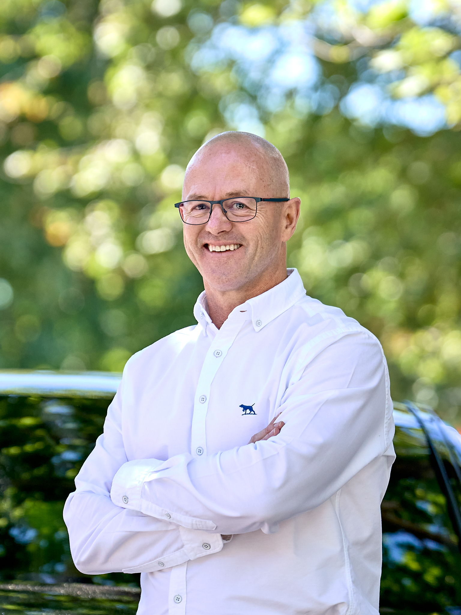 MIchael Clapham wearing a white shirt, arms folded standing in front of a car, with a leafy background.