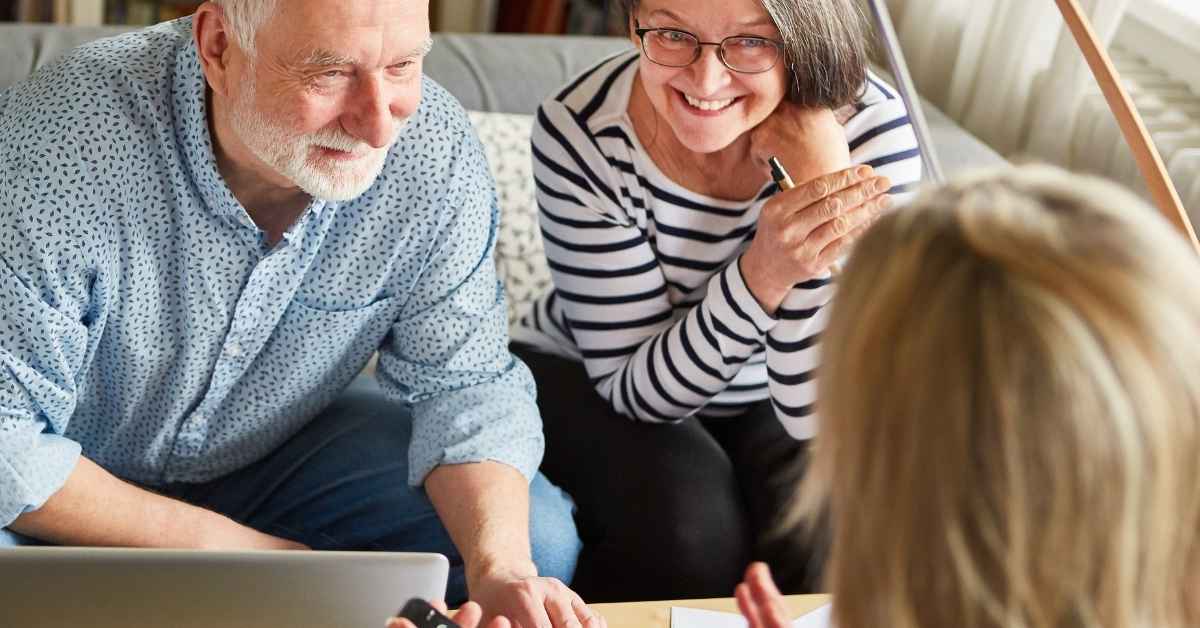 Man with grey hair, blue shirt sits next to his wife on couch, with stripe shirt having a conversation with a financial adviser about retirement.